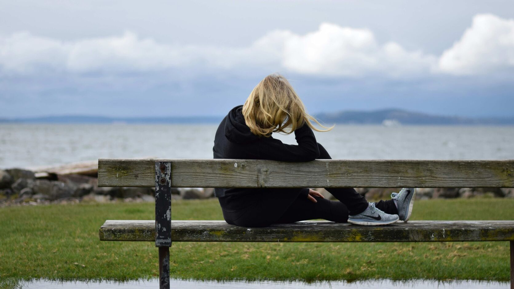 A blonde woman in a black hoodie sits alone on a weathered wooden bench, head bowed, facing a grey, overcast sea.