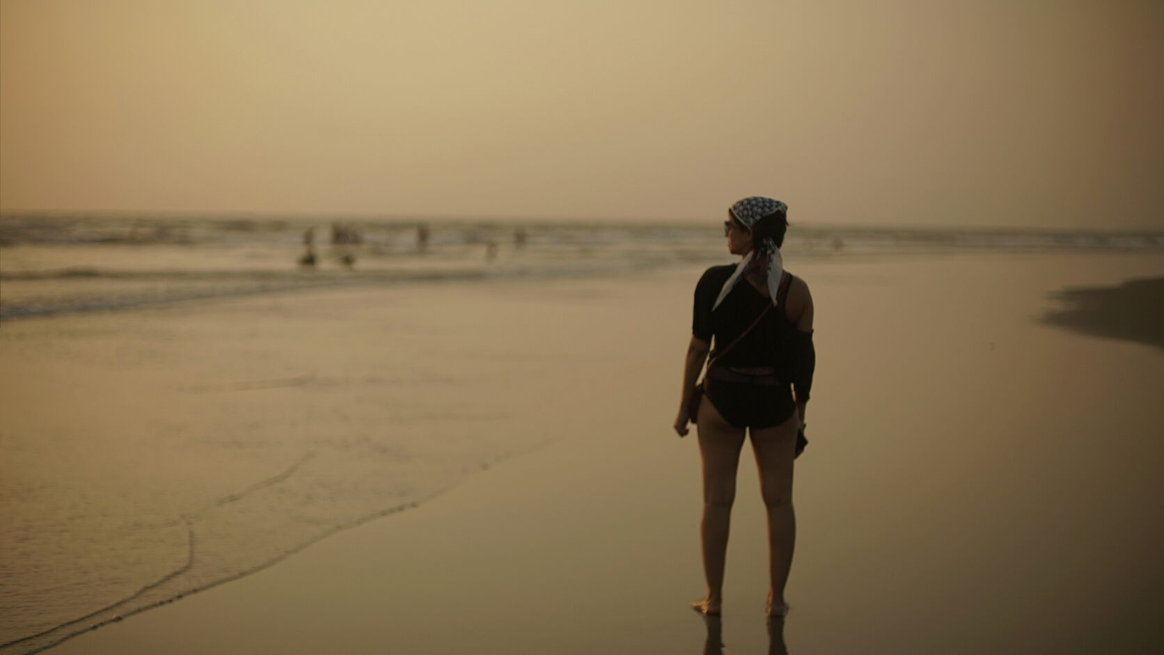 A woman stands alone on a wide beach at sunset, facing the water, the sky washed in soft orange light.
