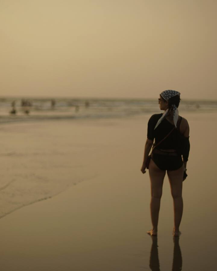 A woman stands alone on a wide beach at sunset, facing the water, the sky washed in soft orange light.
