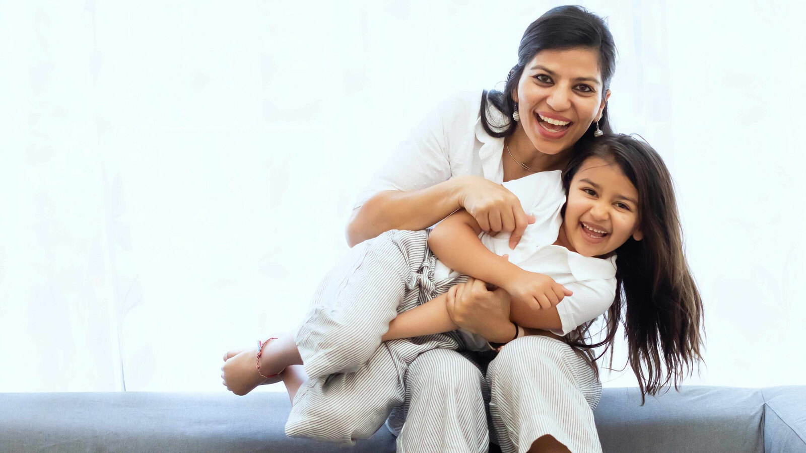 A smiling mother in a white shirt wraps her arms around her laughing young daughter on a couch by a bright window.