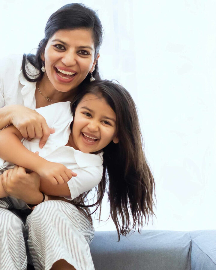 A smiling mother in a white shirt wraps her arms around her laughing young daughter on a couch by a bright window.