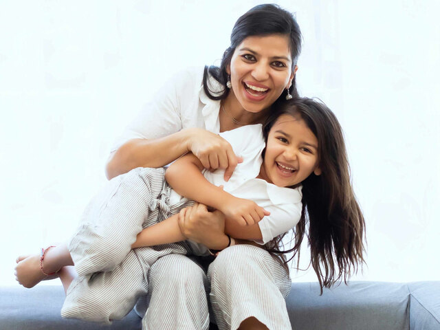 A smiling mother in a white shirt wraps her arms around her laughing young daughter on a couch by a bright window.