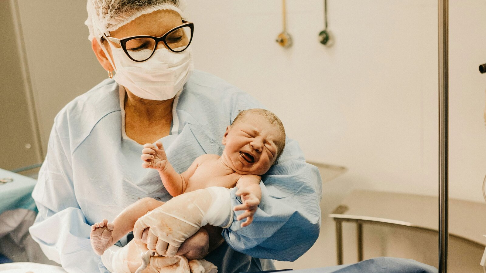 A masked clinician in blue scrubs cradles a crying newborn moments after birth in a hospital delivery room.
