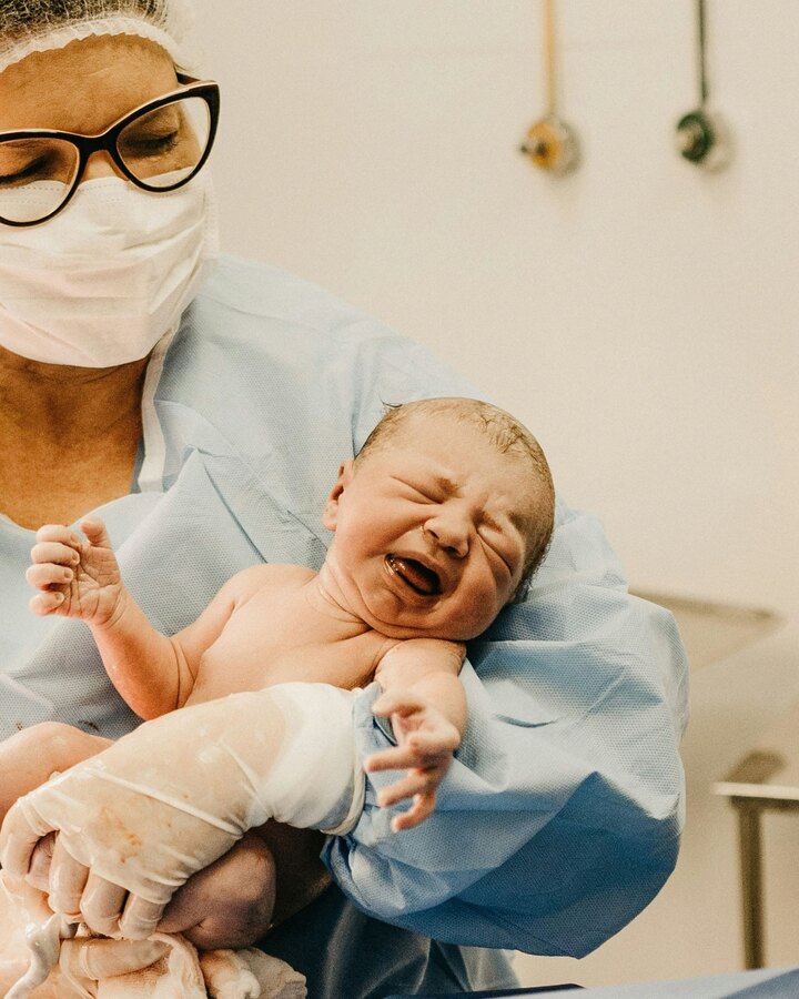 A masked clinician in blue scrubs cradles a crying newborn moments after birth in a hospital delivery room.