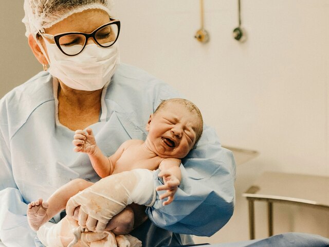 A masked clinician in blue scrubs cradles a crying newborn moments after birth in a hospital delivery room.
