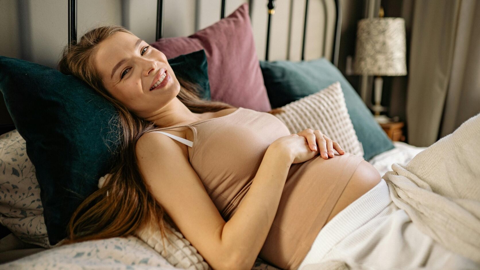 A smiling pregnant woman in a beige top reclines against jewel-toned pillows on a bed, one hand resting on her belly.