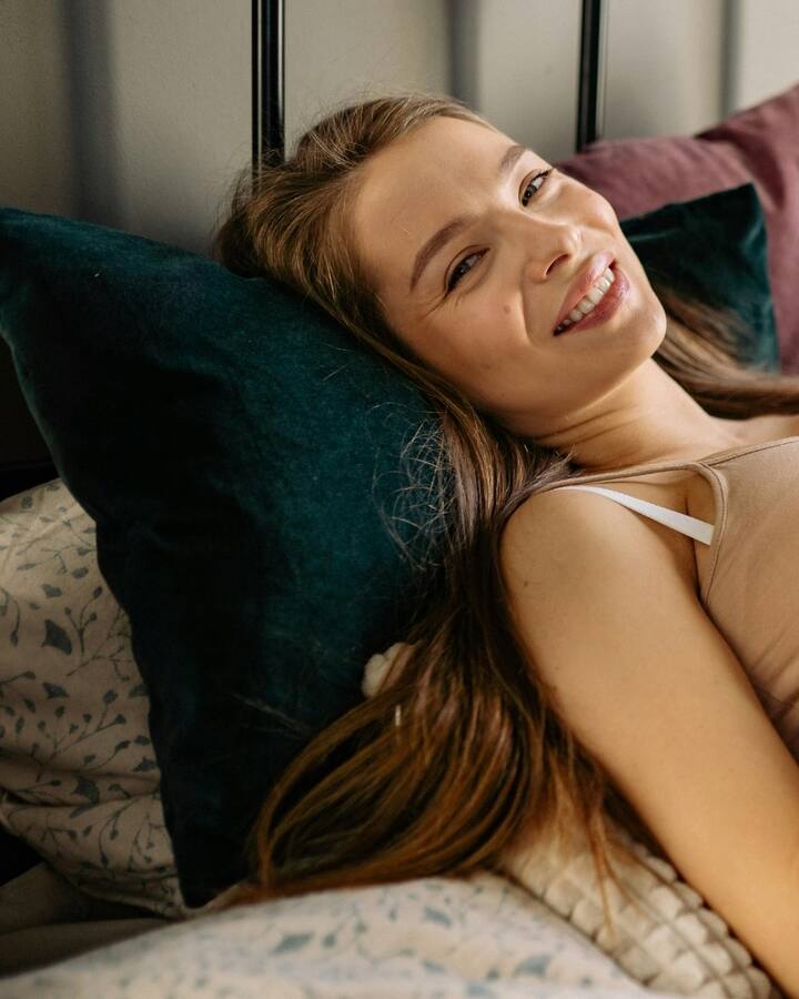 A smiling pregnant woman in a beige top reclines against jewel-toned pillows on a bed, one hand resting on her belly.