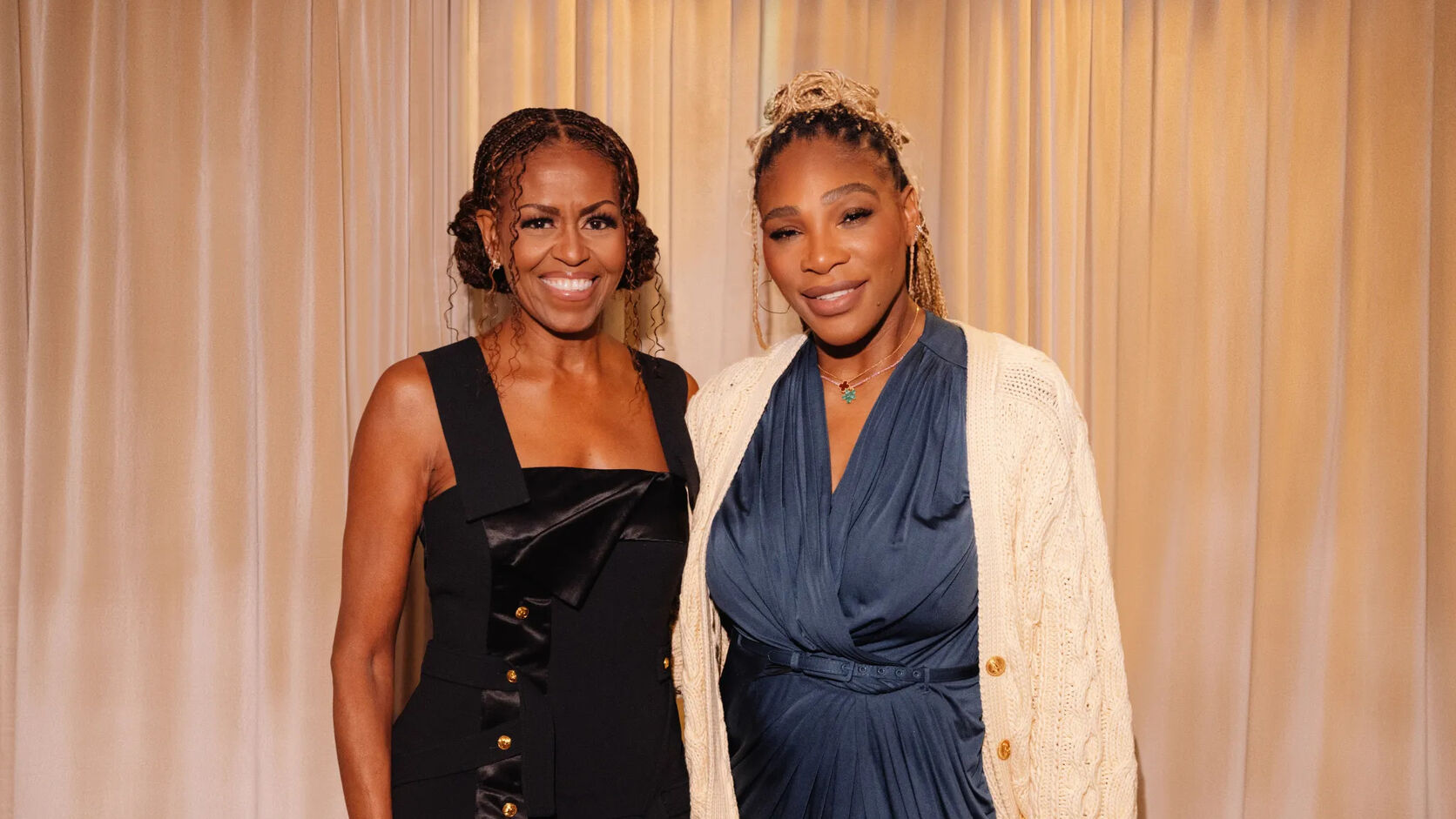 Michelle Obama in a black satin-panelled dress stands beside Serena Williams in a navy dress and cream cable-knit cardigan in front of a softly lit champagne curtain.