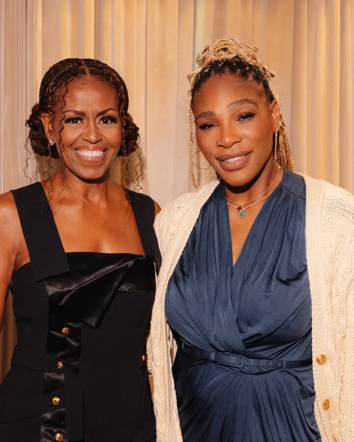 Michelle Obama in a black satin-panelled dress stands beside Serena Williams in a navy dress and cream cable-knit cardigan in front of a softly lit champagne curtain.