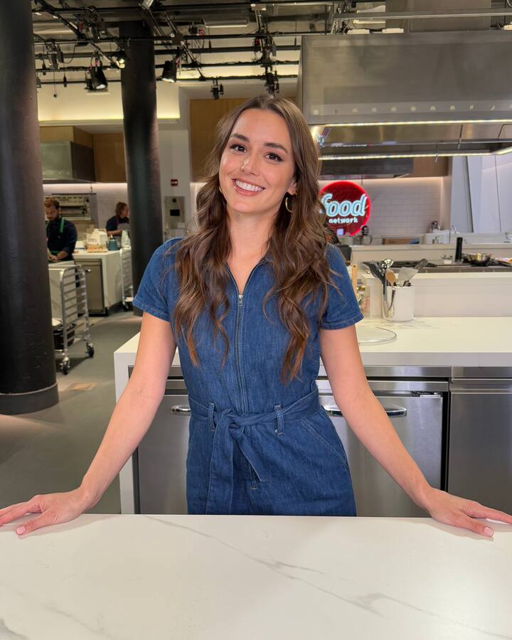 Mika Leon, in a short-sleeved denim jumpsuit and long wavy hair, smiles with both hands resting on a white marble counter in a bright test kitchen.