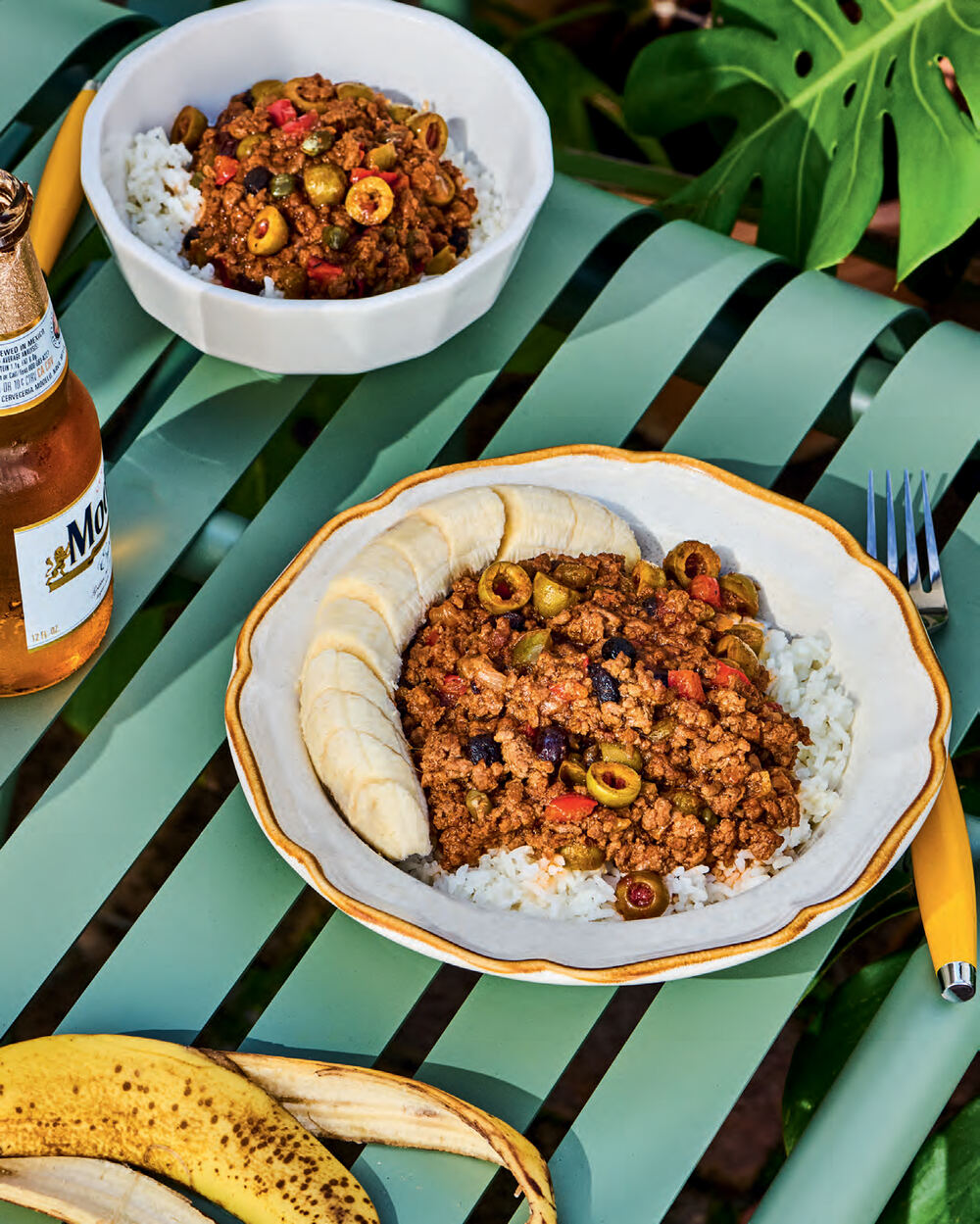 A scalloped plate of picadillo with green olives, capers and raisins over white rice, a sliced ripe banana curved alongside, on a green slatted garden table with a second bowl, a bottle of beer and a banana peel nearby.