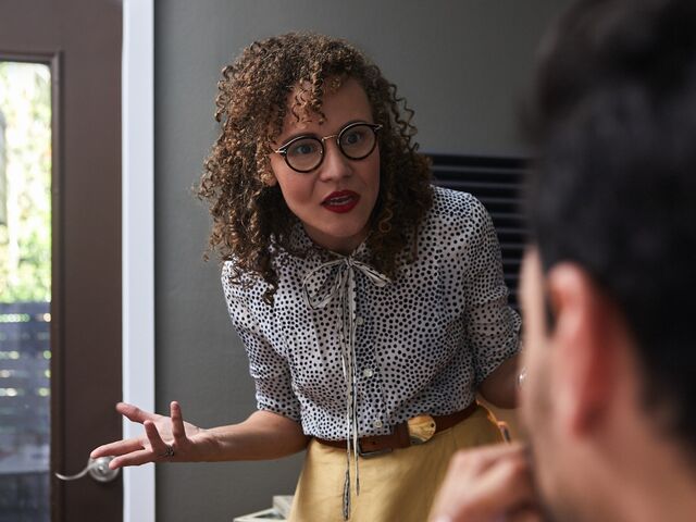 A woman in glasses and a polka-dot blouse mid-sentence, gesturing with frustration toward her partner in a doorway.