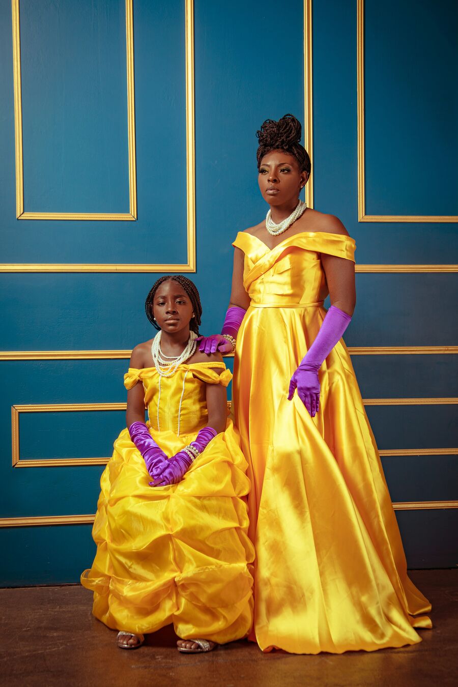 A mother and her daughter in matching yellow satin Belle ball gowns, long purple gloves and layered pearl necklaces pose against a teal wall with gold panel mouldings, the girl seated and the mother standing beside her with a gloved hand on her shoulder.