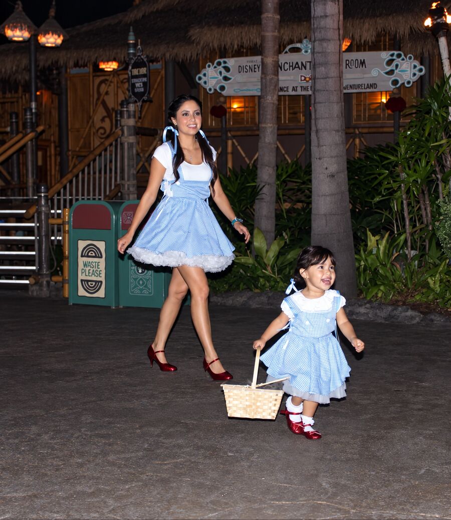 A smiling mother and her toddler daughter in matching blue gingham Dorothy pinafores with white blouses and sparkly red shoes walk along a palm-lined path at night, the little girl laughing as she swings a woven basket.
