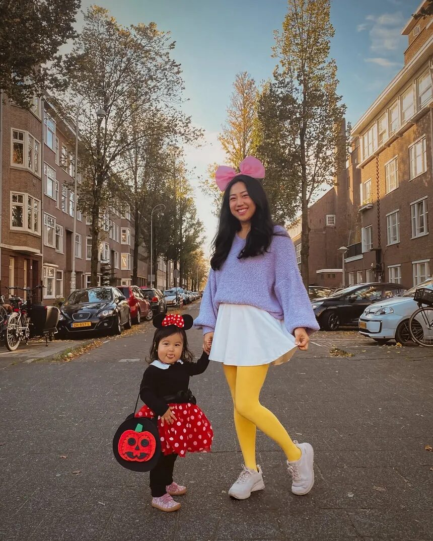 On a tree-lined autumn street of brick apartment buildings, a mother dressed as Daisy Duck, in a big pink bow, lilac sweater, white skirt and yellow tights, holds hands with her toddler dressed as Minnie Mouse, in mouse ears and a red polka-dot skirt, carrying a jack-o&rsquo;-lantern bag and sticking out her tongue.