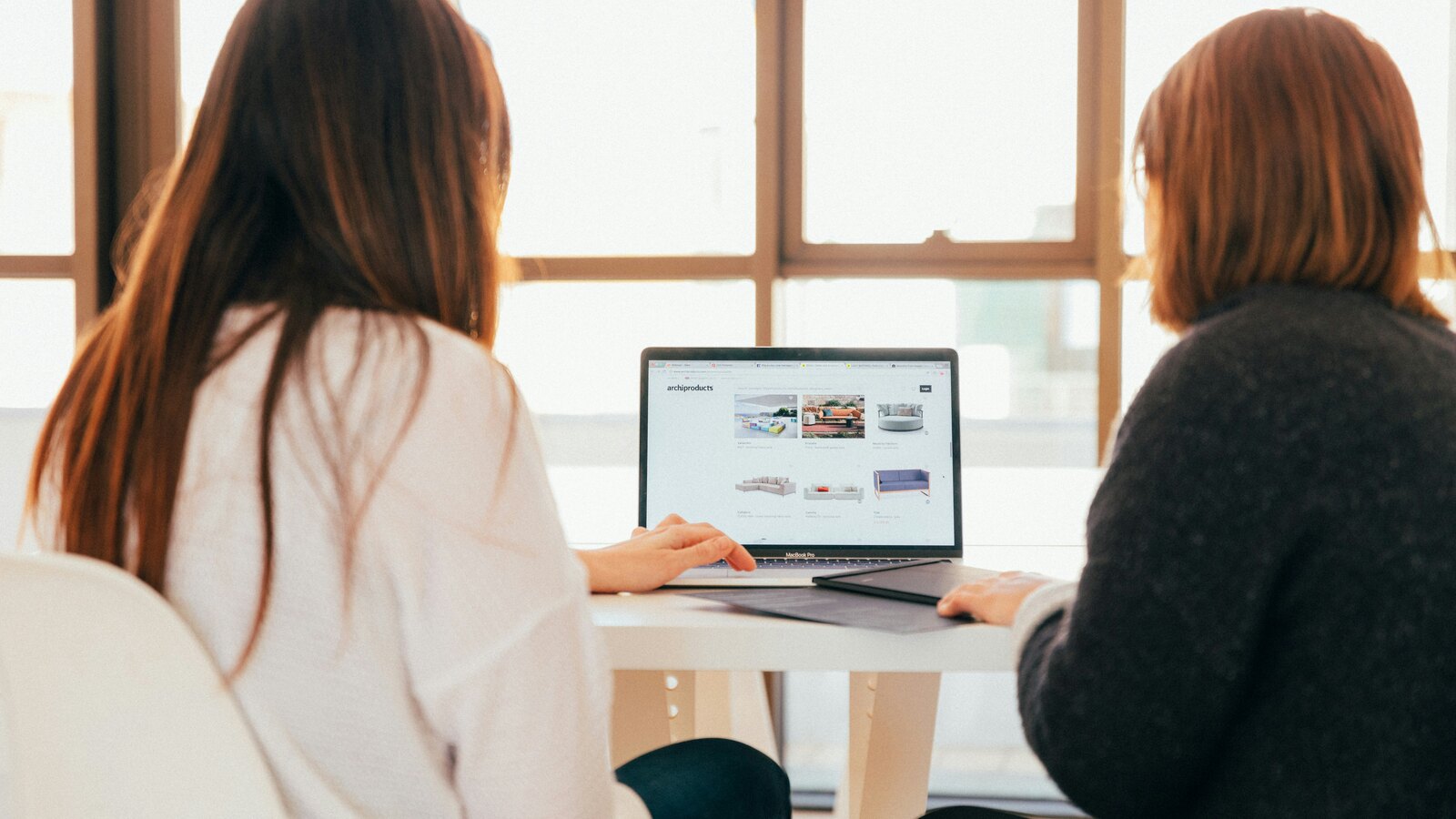 Two women sit side by side at a white table in a sunlit room, looking together at a laptop showing an e-commerce storefront.