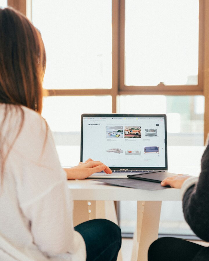 Two women sit side by side at a white table in a sunlit room, looking together at a laptop showing an e-commerce storefront.