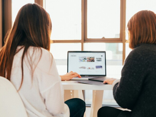 Two women sit side by side at a white table in a sunlit room, looking together at a laptop showing an e-commerce storefront.