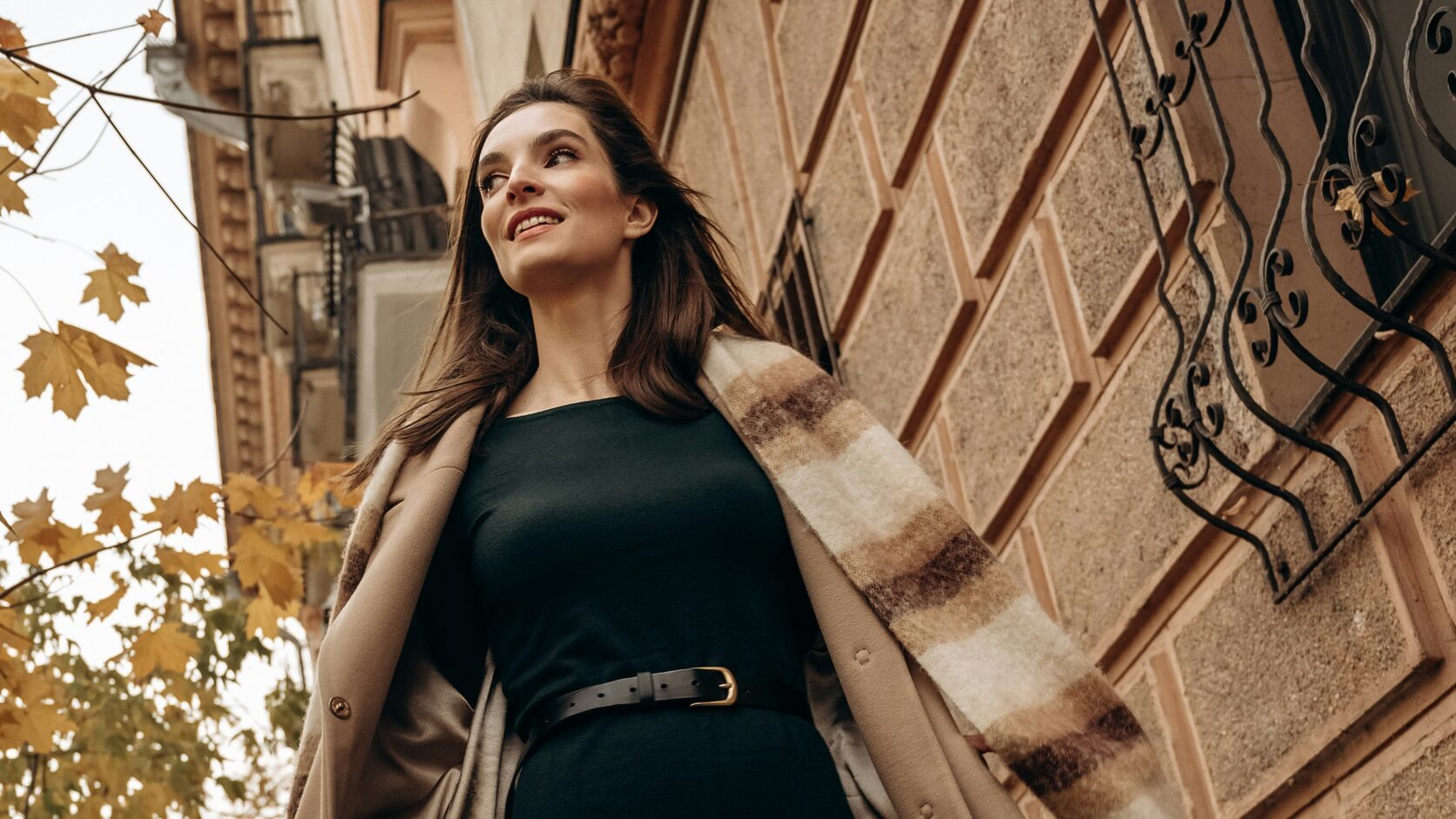 A woman walks down a city street in a camel wool coat over a black belted dress, a cream and brown plaid scarf flying behind her, yellow maple leaves overhead.