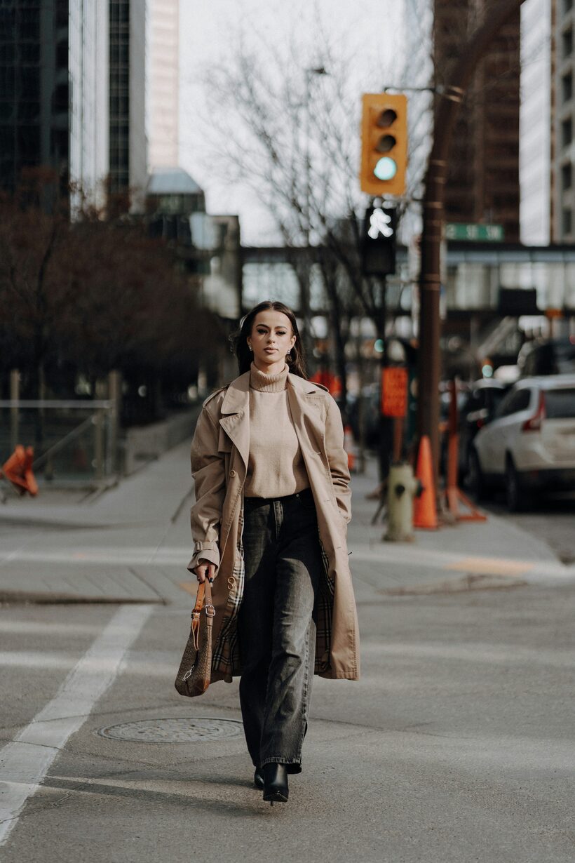 A woman crosses a downtown intersection in a camel trench coat over a beige turtleneck, wide dark jeans, and black heeled boots, a brown shoulder bag on her arm.