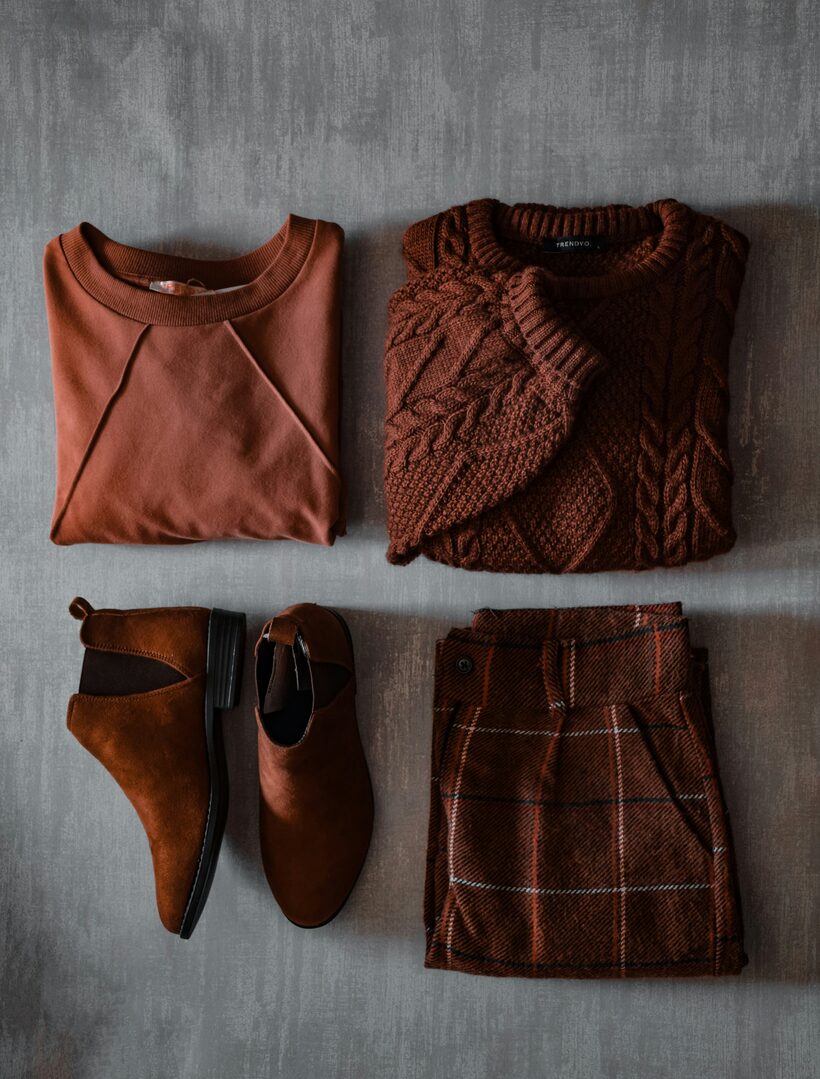 A flat lay on grey concrete: a rust sweatshirt, a chocolate cable-knit sweater, brown suede ankle boots, and a rust plaid skirt.