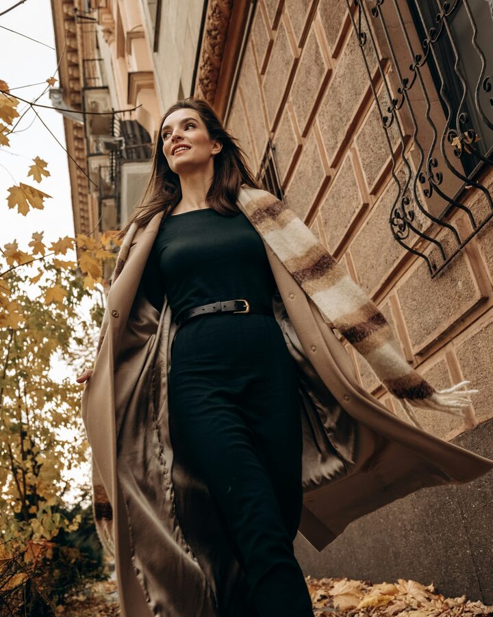 A woman walks down a city street in a camel wool coat over a black belted dress, a cream and brown plaid scarf flying behind her, yellow maple leaves overhead.