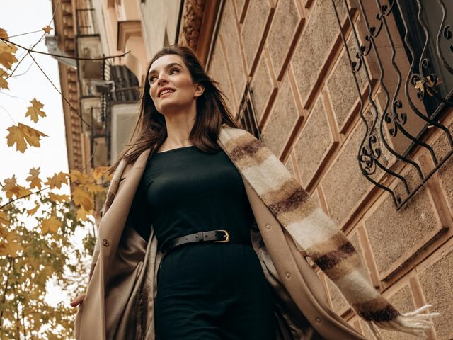A woman walks down a city street in a camel wool coat over a black belted dress, a cream and brown plaid scarf flying behind her, yellow maple leaves overhead.