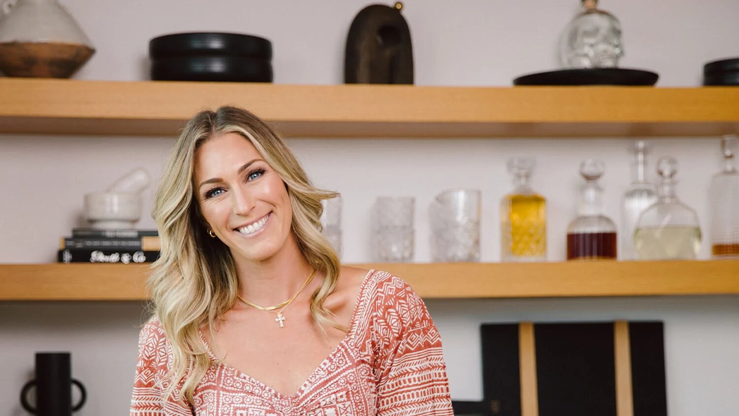 Dr. Morgan Cutlip smiles in a rust-and-cream printed dress, standing in a bright kitchen in front of open wood shelves.