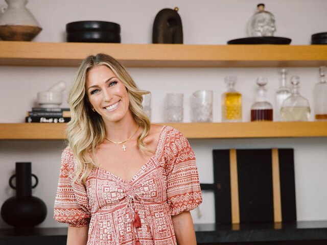 Dr. Morgan Cutlip smiles in a rust-and-cream printed dress, standing in a bright kitchen in front of open wood shelves.