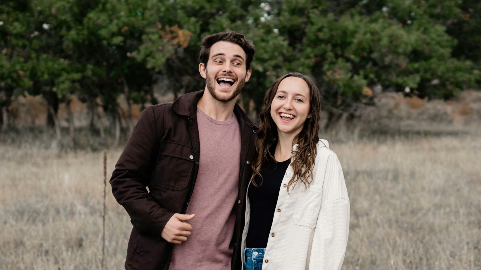 A young man in a brown jacket and a young woman in a cream corduroy shirt stand side by side in a field of dry grass, both laughing, trees behind them.