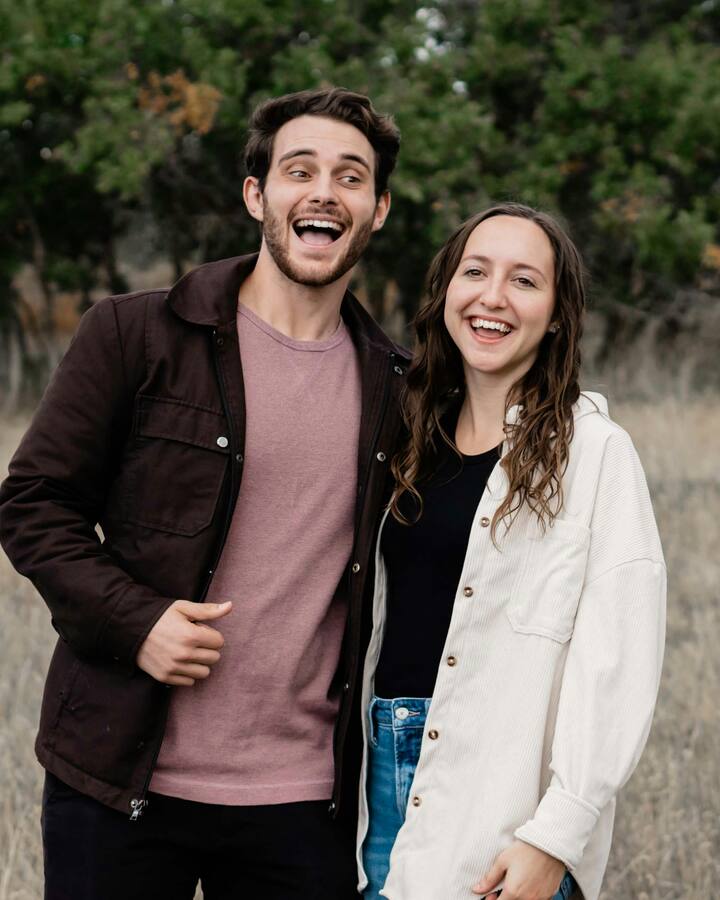 A young man in a brown jacket and a young woman in a cream corduroy shirt stand side by side in a field of dry grass, both laughing, trees behind them.
