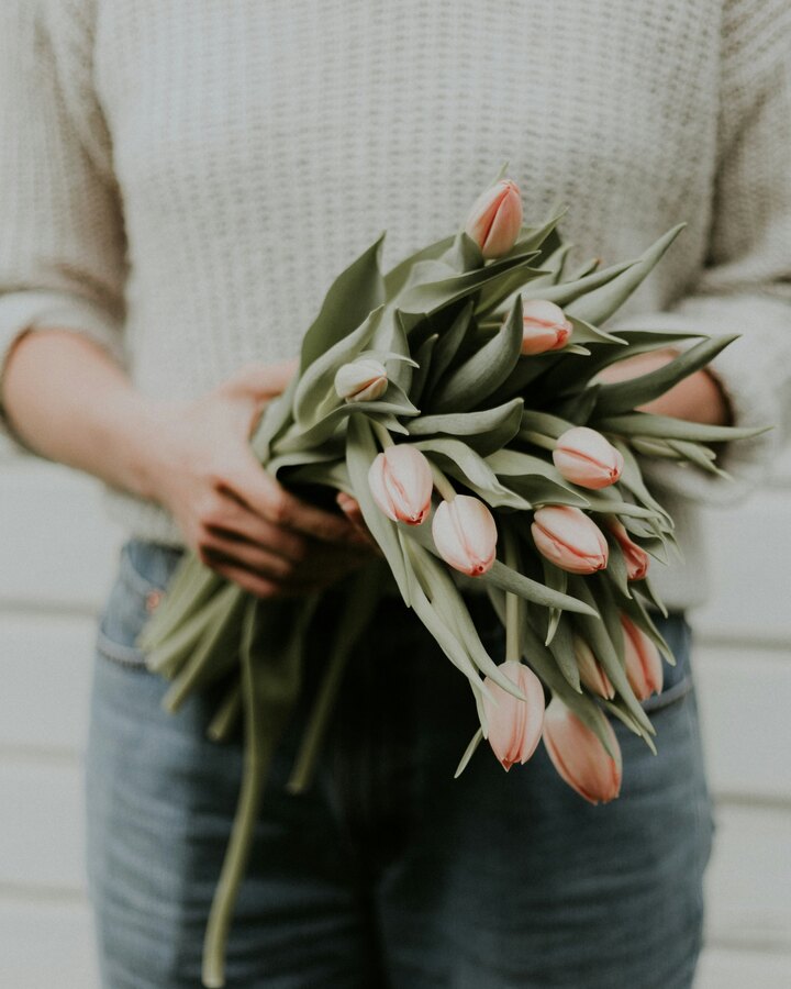 A woman in a cream knit sweater and jeans holds a loose armful of blush pink tulips against a white clapboard wall.