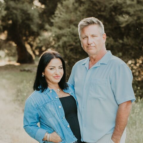 Nellie Akalp and her husband Phil stand together on a gravel path lined with olive trees, she in a denim jacket over a black maxi dress, he in a light blue shirt and cream trousers.