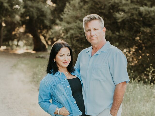 Nellie Akalp and her husband Phil stand together on a gravel path lined with olive trees, she in a denim jacket over a black maxi dress, he in a light blue shirt.