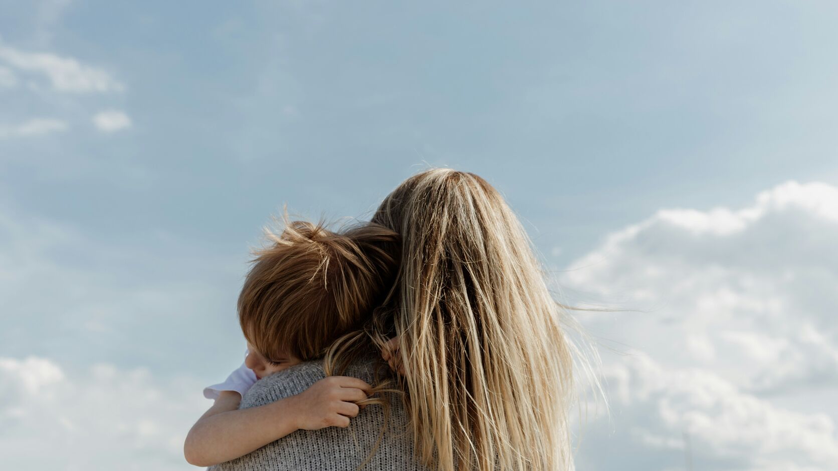 A mother with windblown blonde hair holds her young child close against her shoulder under a wide, soft blue sky.