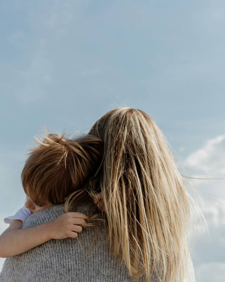 A mother with windblown blonde hair holds her young child close against her shoulder under a wide, soft blue sky.