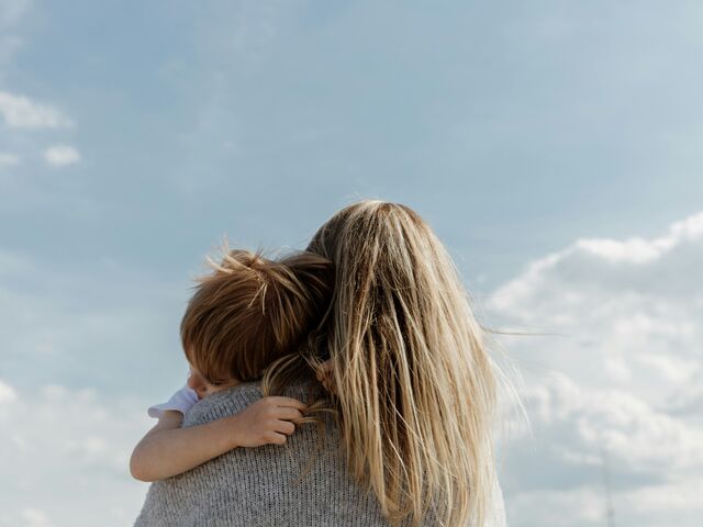 A mother with windblown blonde hair holds her young child close against her shoulder under a soft blue sky.