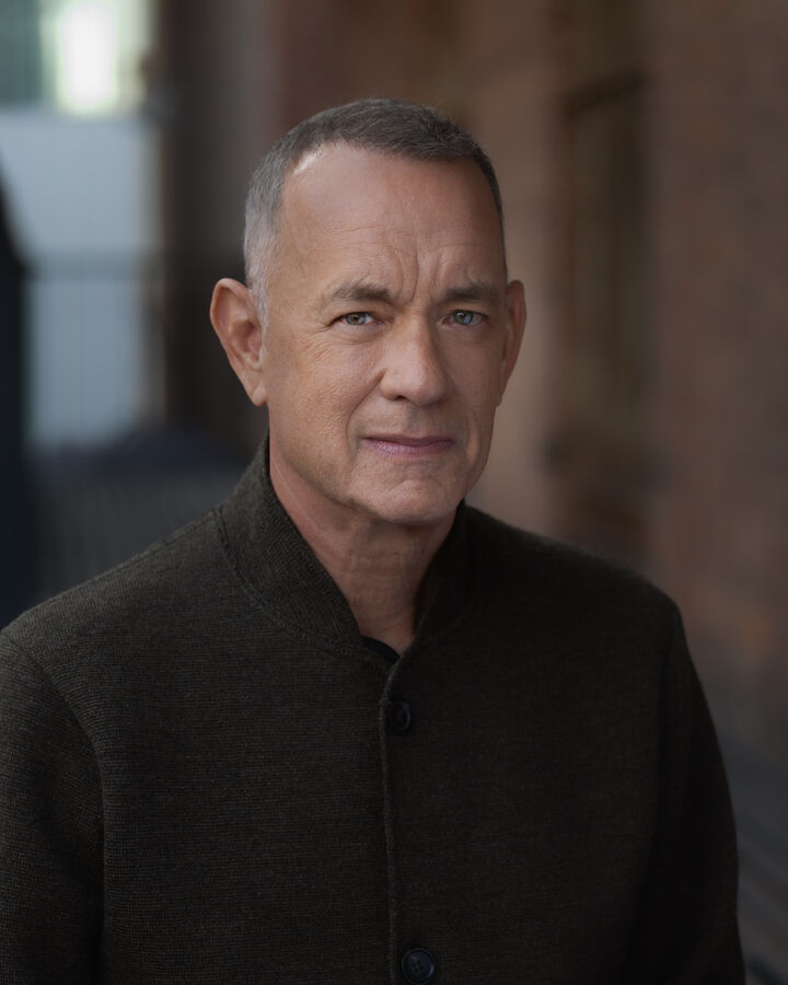 Tom Hanks in a dark knit jacket, photographed against a softly blurred brick wall.