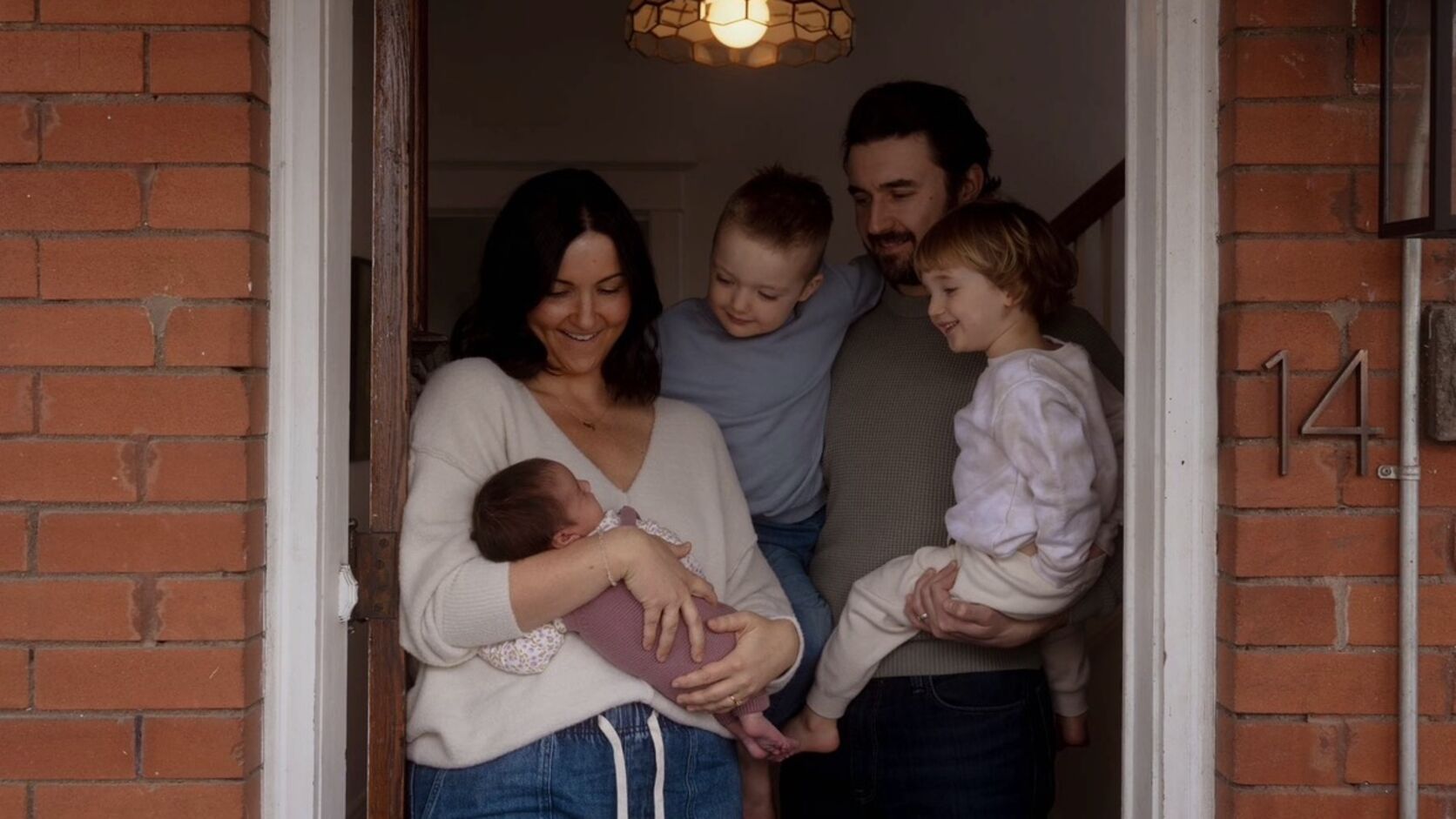 Page Cunningham stands in the front doorway of a red-brick house in a cream sweater, cradling her newborn daughter, while her husband holds their two older children beside her.