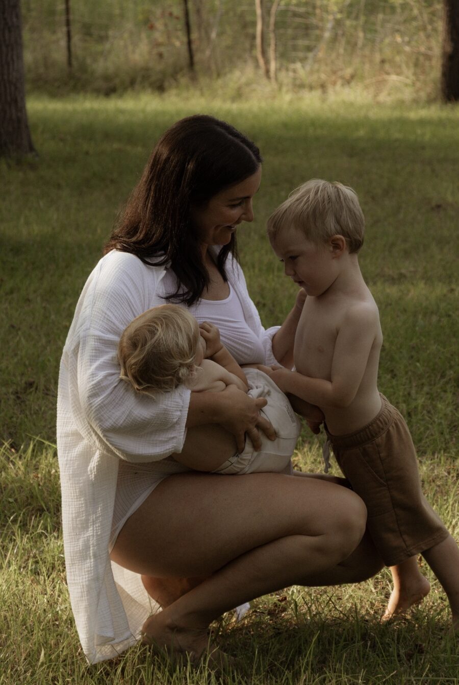 Page Cunningham crouches barefoot on a sunlit lawn in a white gauze shirt, nursing her baby while her toddler son leans in against her.