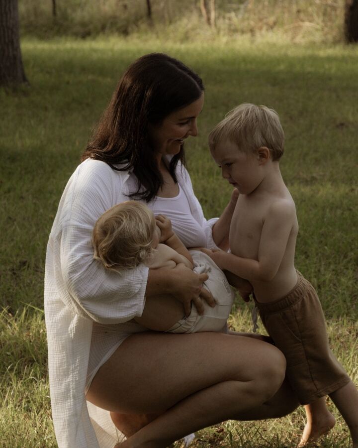 Paige Cunningham crouches barefoot on a sunlit lawn in a white gauze shirt, nursing her baby while her toddler son leans in against her.
