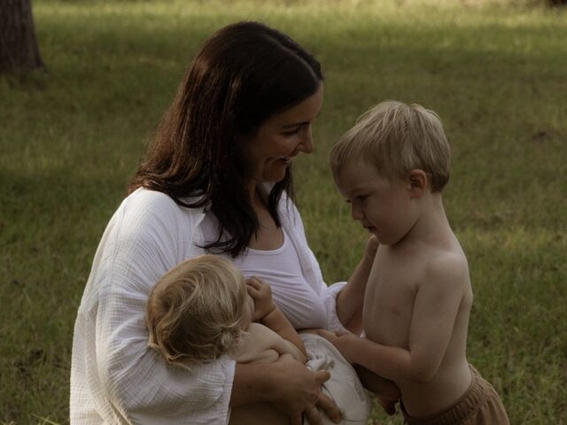 Paige Cunningham nurses her baby on a sunlit lawn while her toddler son leans in against her.
