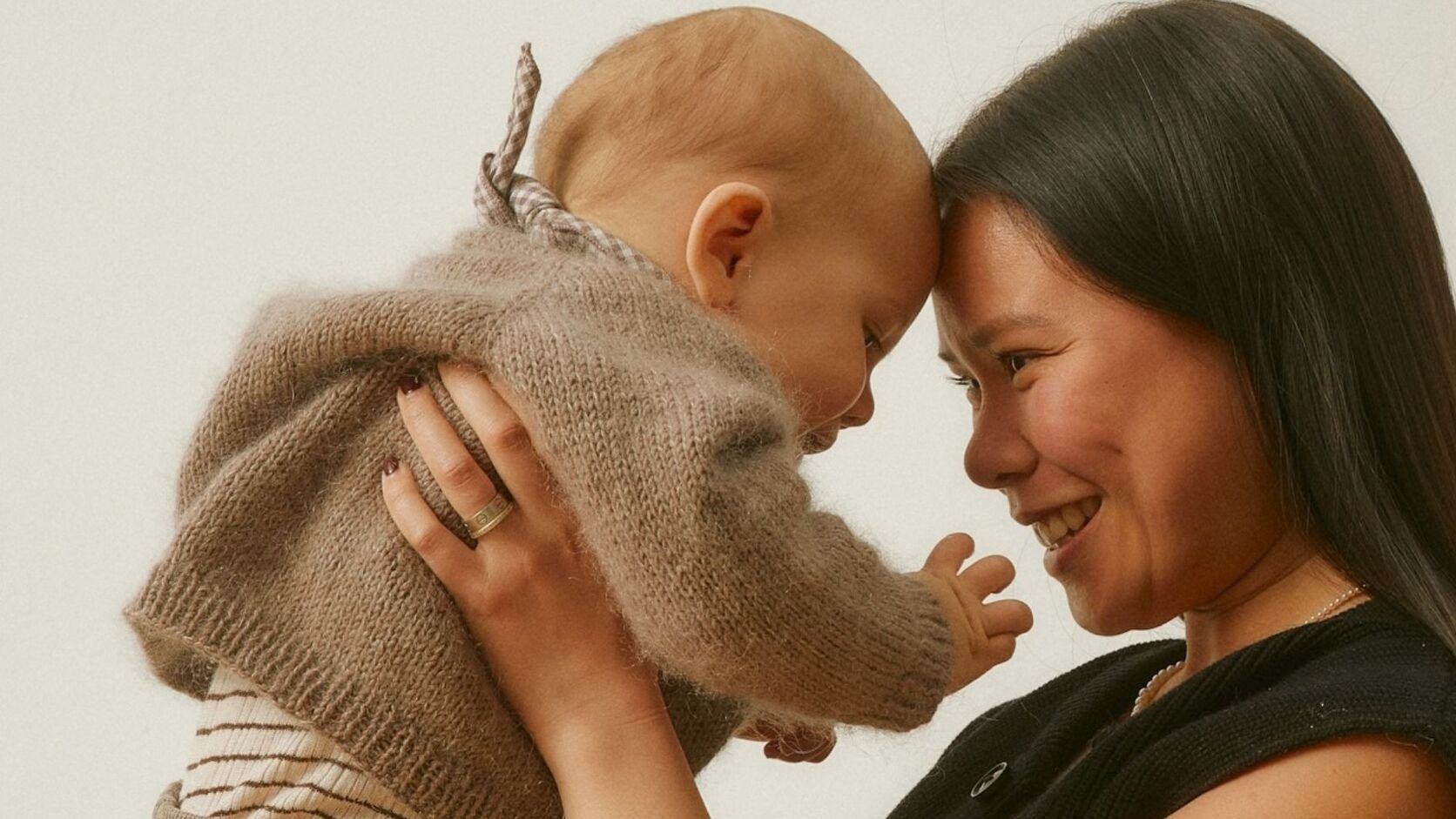 A smiling mother holds her baby close, foreheads touching, against a soft cream background.