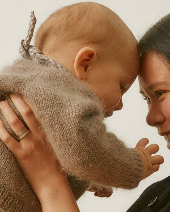 A smiling mother holds her baby close, foreheads touching, against a soft cream background.