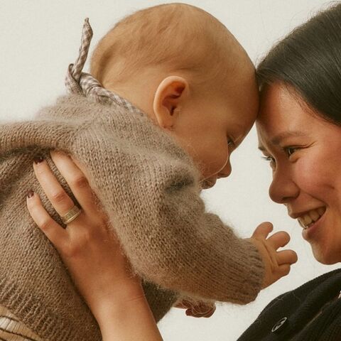 A smiling mother holds her baby close, foreheads touching, against a soft cream background.