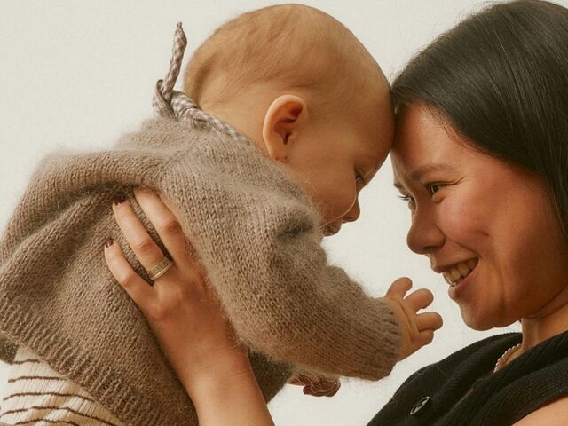 A smiling mother holds her baby close, foreheads touching, against a soft cream background.