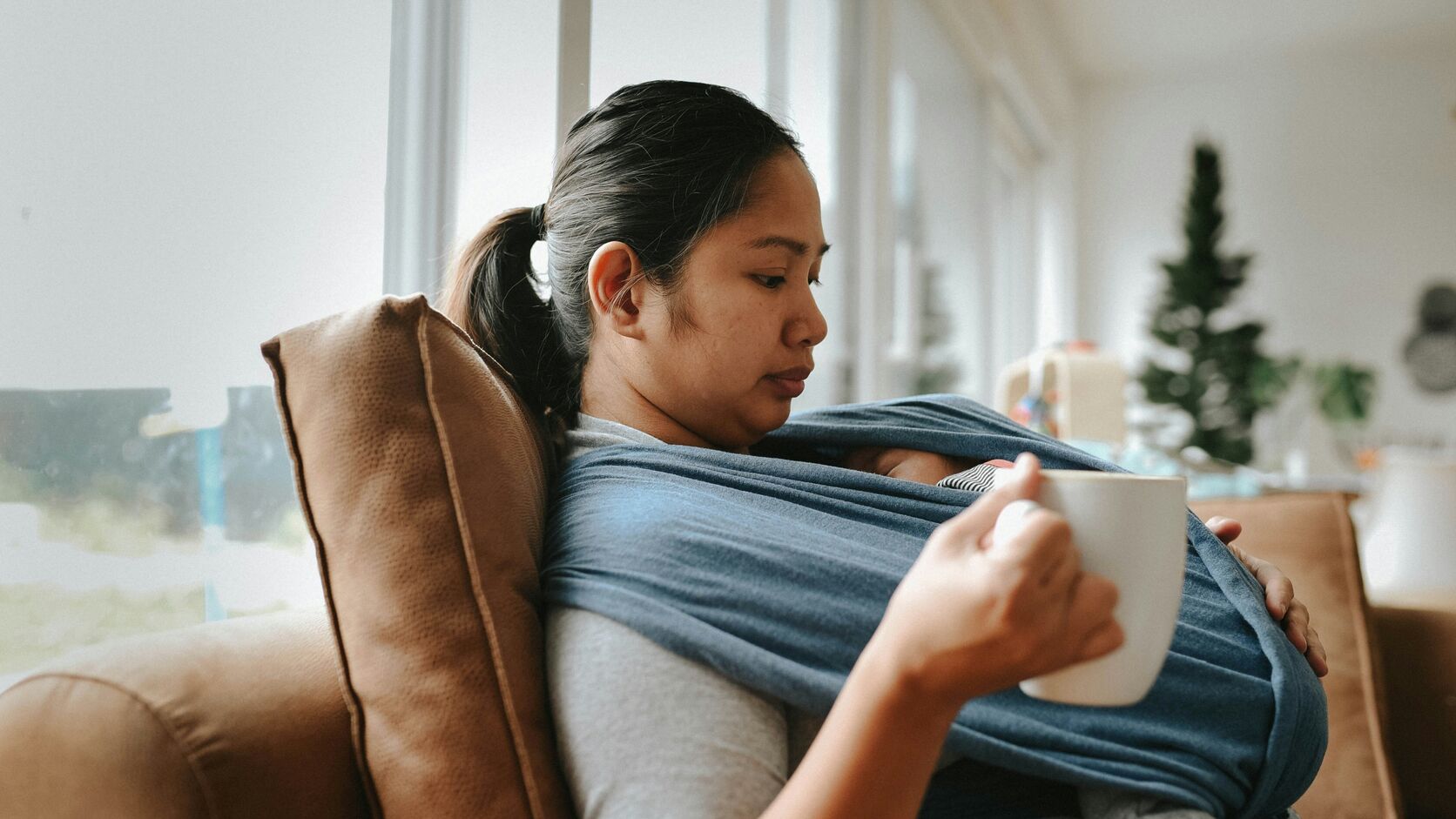 A mother sits back on a sofa in soft window light holding a mug, her newborn asleep against her chest in a blue wrap, her eyes lowered.