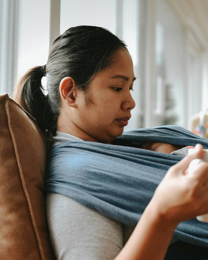 A mother sits back on a sofa in soft window light holding a mug, her newborn asleep against her chest in a blue wrap, her eyes lowered.
