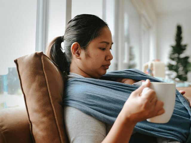 A mother sits back on a sofa in soft window light holding a mug, her newborn asleep against her chest in a blue wrap, her eyes lowered.