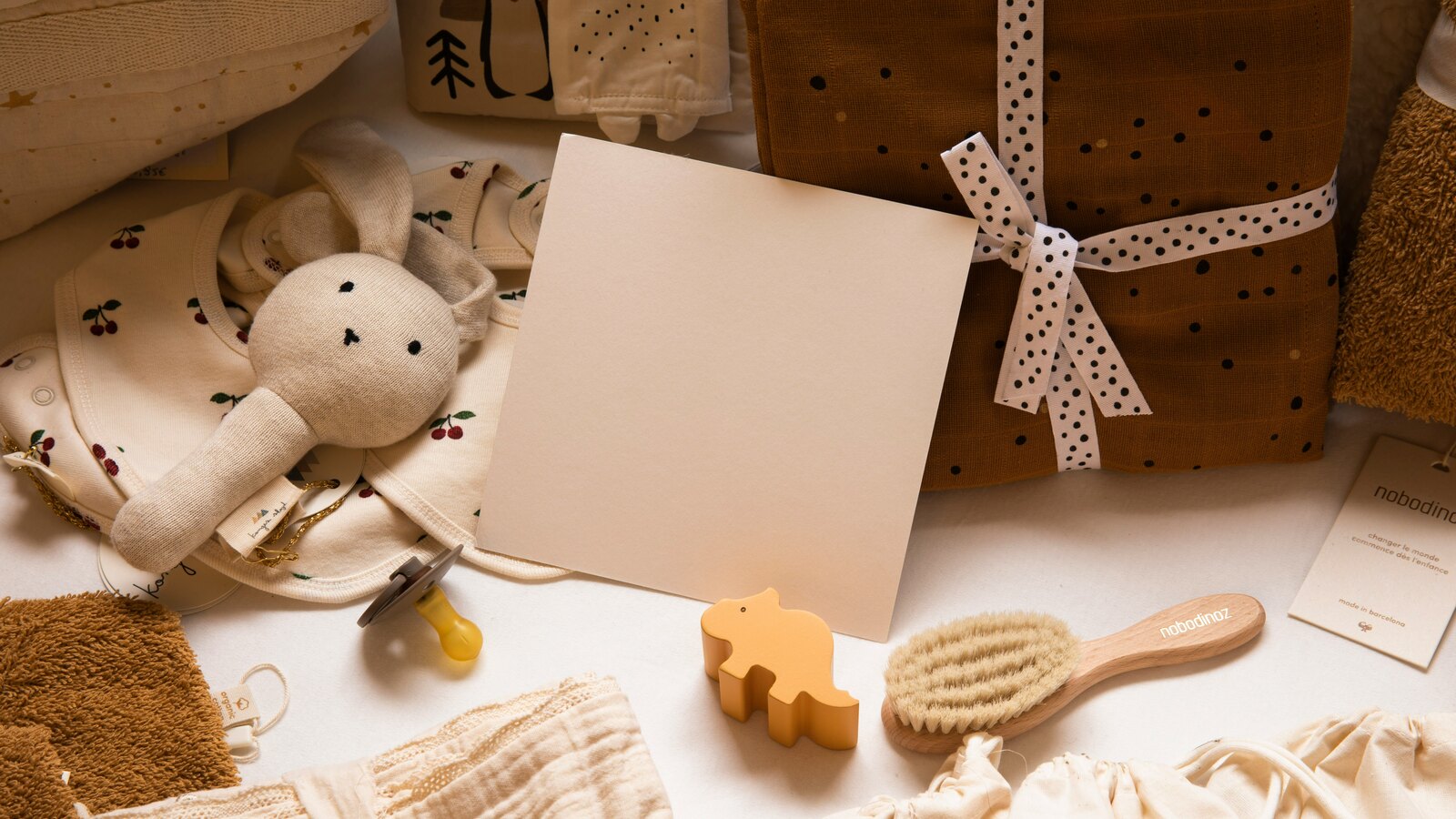 A neutral-toned flat-lay of baby items — a cream knit bunny rattle, a cherry-print onesie, a wooden brush, muslin wraps tied with polka-dot ribbon, and a blank cream card.